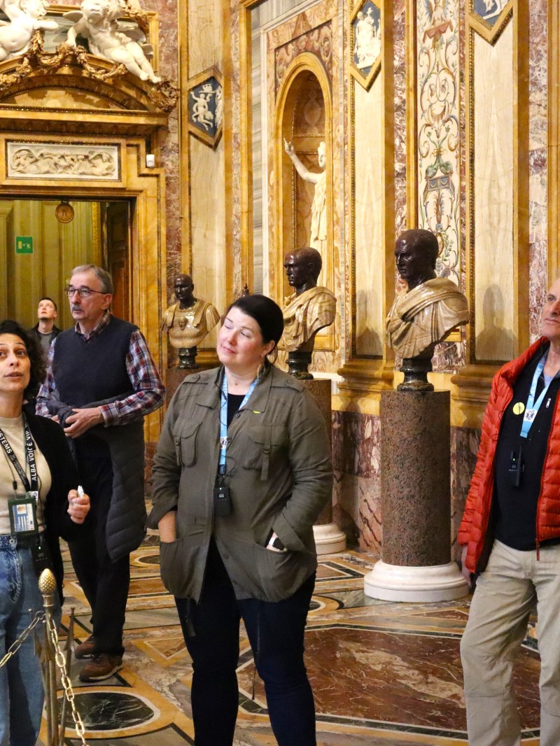 Tour guide showing group of six people around a museum with statues and ornate decor.