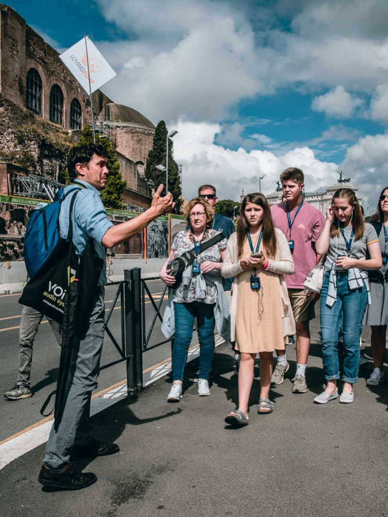 Tour guide with a group of people on a city street near historical ruins under a partly cloudy sky.