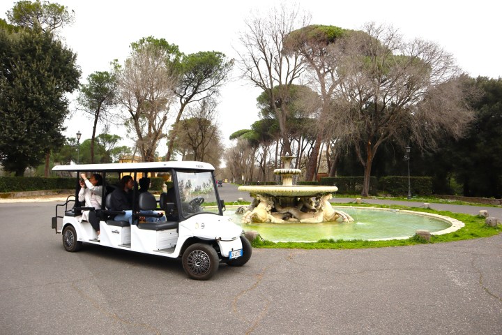 People on a white golf cart ride past a decorative fountain in a park setting.