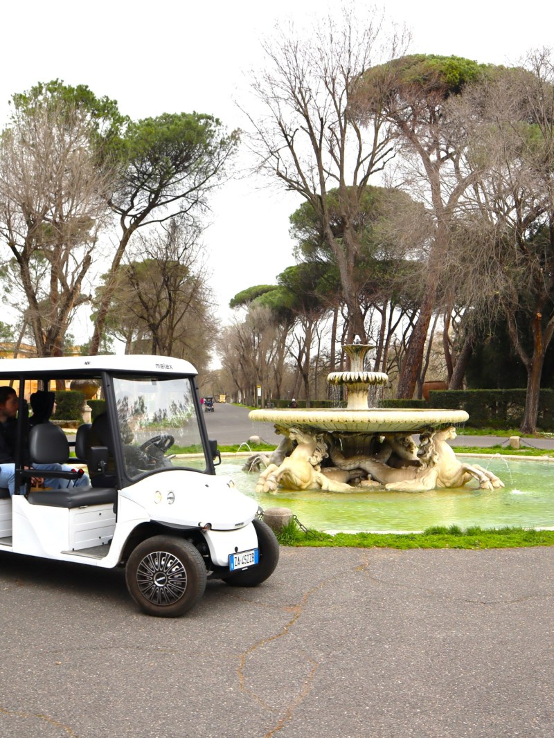 People on a white golf cart ride past a decorative fountain in a park setting.