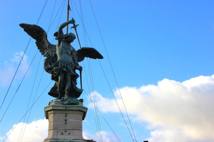 Angel statue with sword and wings, standing on a pedestal against a blue sky with clouds.