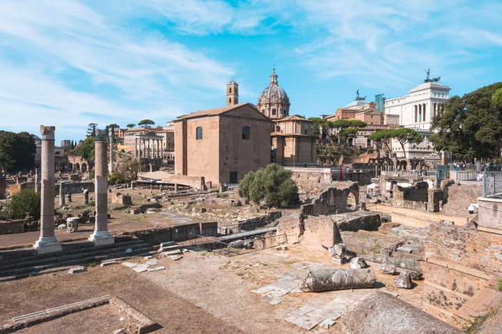 Ancient Roman ruins with columns and distant historic buildings under a blue sky.