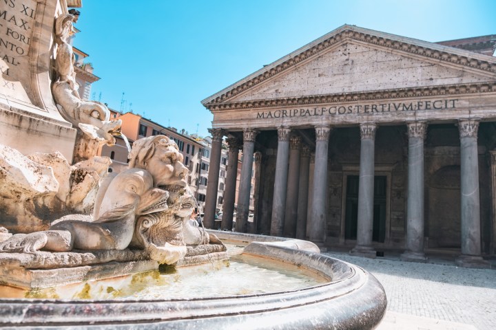 Fountain and columns in front of an ancient Roman building with Latin inscription.