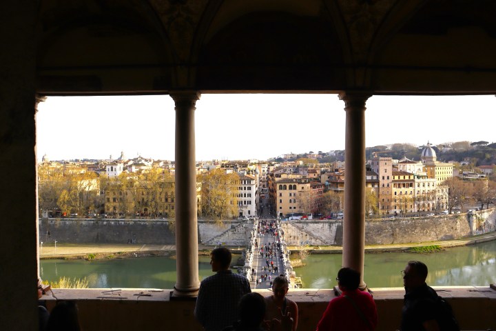 People view Roman cityscape from shaded terrace overlooking a bridge and river.