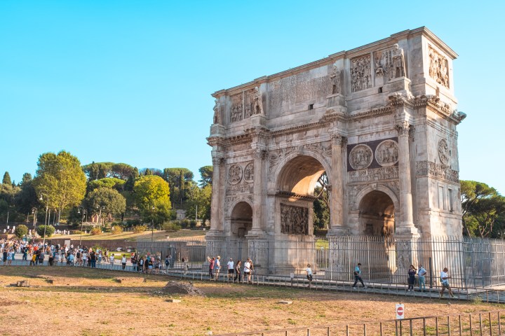 A large ancient stone arch with intricate carvings, surrounded by tourists and trees under a clear blue sky.
