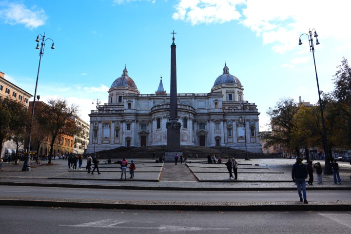 People walking near a large historic building with domes and an obelisk in front.
