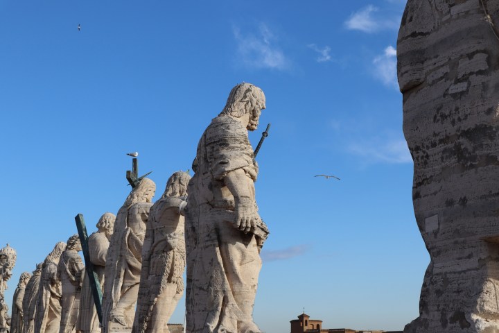 Row of stone statues with a blue sky and flying birds in the background.